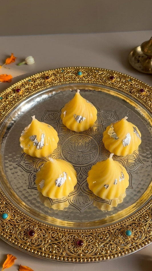 Five yellow marigold flower-shaped sweets decorated with silver foil, arranged on an ornate gold-toned metal tray with floral accents on a neutral surface.