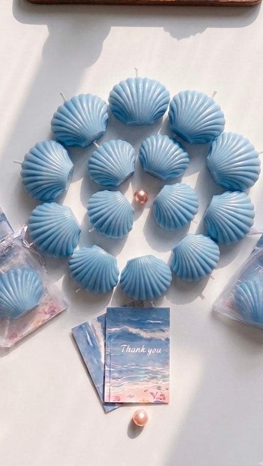 A set of light blue seashell-shaped candles arranged in a circle on a white surface, with a small pearl-like bead and a printed card featuring a beach scene and the words 'Thank you'.