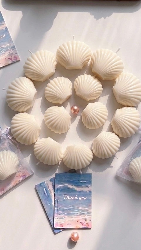 A circular arrangement of white shell-shaped candles on a light surface, with a few matching decorative cards and pearls placed around them.