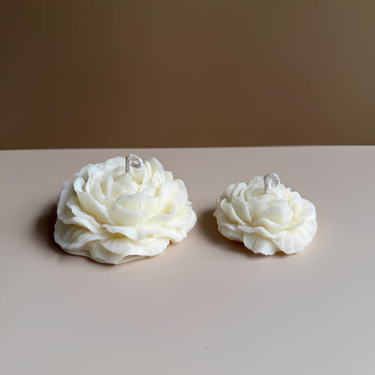 Two white flower-shaped candles on a beige surface with a brown background.Peony Bloom Grandé sculptural peony soy wax candle for luxury home décor and gifting
